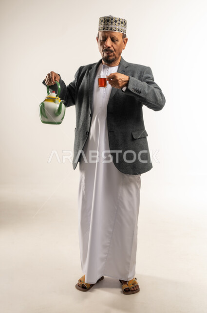 مقاومة الشعور بالبرد، رفع حرارة الجسم وتدفئته، احتساء مشروب ساخن، الصقيع والأجواء الشتوية الباردة، ارتداء ملابس الشتاء الثقيلة، بورتريه لرجل عربي خليجي عماني يرتدي الكمه والمعطف الشتوي يقوم بصب الشاي من الابريق، الجسم بالطول الكامل، خلفية بيضاء