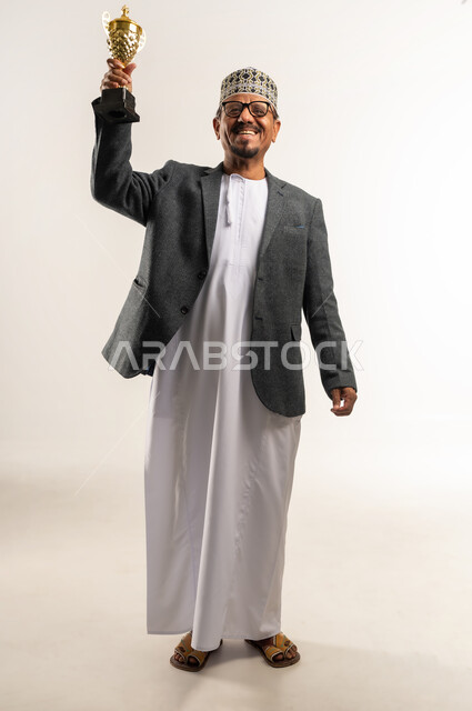 الحصول على المركز الأول في المسابقة، التتويج بالبطولة والفوز، النظر إلى الكاميرا بإيماءات السعادة والسرور، بورتريه لرجل عربي خليجي عماني يرتدي الدشداشه و الكمه والمعطف الشتوي يحمل بيده الكأس الذهبية بتعابير الحماس، الجسم بالطول الكامل, خلفية بيضاء