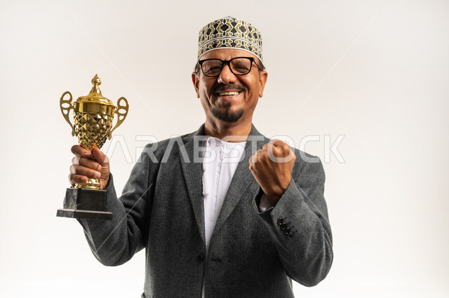 رفع اليد في الهواء بتعابير الحماس، الحصول على المركز الأول في المسابقة، التتويج بالبطولة والفوز، النظر إلى الكاميرا بإيماءات السعادة، بورتريه مقرب لرجل عربي خليجي عماني يرتدي الدشداشه و الكمه والمعطف الشتوي يحمل بيده الكأس الذهبية، خلفية بيضاء
