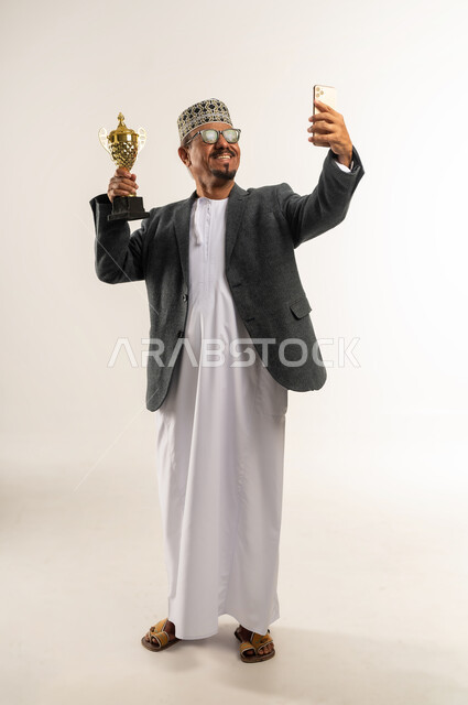 التقاط صورة تذكارية مع الكأس الذهبية, استخدام التقنيات التكنولوجية الحديثة والمتطورة,  إيماءات السعادة و السرور بالفوز بالبطولة، بورتريه لرجل عربي خليجي عماني يرتدي الدشداشه و الكمه والمعطف الشتوي يأخذ لقطة سيلفي عبر الجوال, الجسم بالطول الكامل, خلفية بيضاء