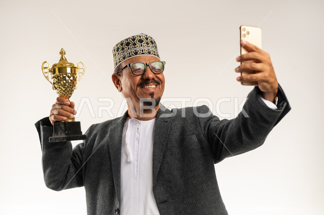 إيماءات السعادة و السرور بالفوز بالبطولة، التقاط صورة تذكارية مع الكأس الذهبية, استخدام التقنيات التكنولوجية الحديثة والمتطورة,  بورتريه مقرب لرجل عربي خليجي عماني يرتدي الدشداشه و الكمه والمعطف الشتوي يأخذ لقطة سيلفي عبر الجوال, خلفية بيضاء