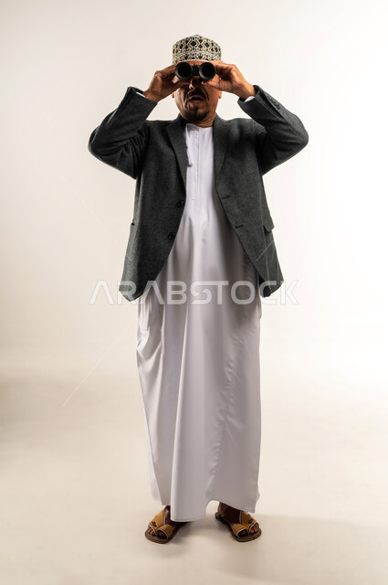 Goals and future plans, clarifying and bringing the vision closer through a telescope, love of exploration and adventure, seeing something or a distant place, portrait of an Arab Gulf Omani man wearing a dishdasha, a sleeve and a winter coat, holding binoculars in his hands and looking through them, full-length body, white background