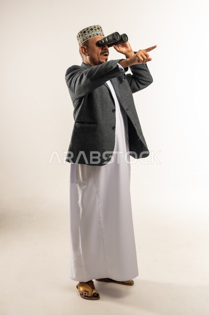 توضيح الرؤية وتقريبها عبر التلسكوب, الإشارة باصبع السبابة إلى مكان ما بعيد، حب الاستكشاف و المغامرة, بورتريه من الجانب لرجل عربي خليجي عماني يرتدي الدشداشة والكمه والمعطف الشتوي يحمل بيده منظار ثنائي العينين وينظر من خلاله، الجسم بالطول الكامل, خلفية بيضاء