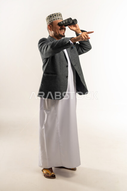 الإشارة باصبع السبابة إلى مكان ما بعيد، توضيح الرؤية وتقريبها عبر التلسكوب, حب الاستكشاف و المغامرة, بورتريه من الجانب لرجل عربي خليجي عماني يرتدي الدشداشة والكمه والمعطف الشتوي يحمل بيده منظار ثنائي العينين وينظر من خلاله، الجسم بالطول الكامل, خلفية بيضاء