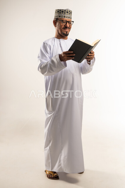 Love of reading and studying, spending free time learning and educating, portrait of an Arab Gulf Omani man wearing a dishdasha, a turban and glasses, holding a book in his hand and reading from it with gestures of concentration, interest in browsing useful books, increasing intellectual stock, full-length body, white background