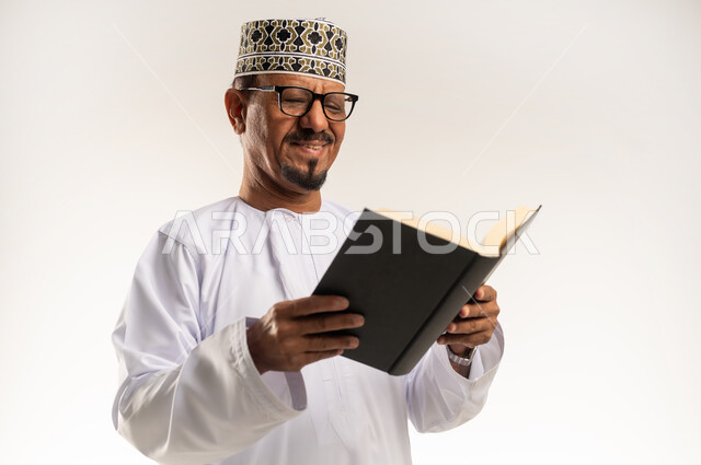 تنمية وزيادة الذخيرة الفكرية، الاهتمام بتصفح الكتب المفيدة، تمضية أوقات الفراغ بالتعلم والتثقيف، حب المطالعة والقراءة، بورتريه مقرب لرجل عربي خليجي عماني يرتدي الدشداشه والكمه والنظارة الطبية يحمل بيده كتاب يقرأ منه بإيماءات التركيز، خلفية بيضاء