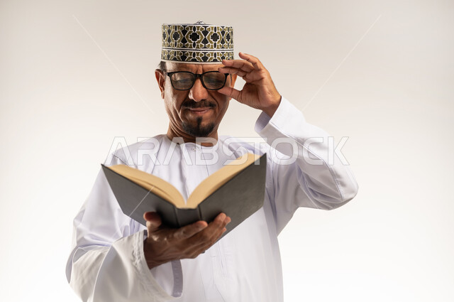 Interest in browsing useful books, spending free time learning and educating, love of reading, close-up portrait of an Arab Gulf Omani man wearing a dishdasha, a turban and glasses, holding a book in his hand and reading from it with gestures of concentration, developing and increasing intellectual stock, white background