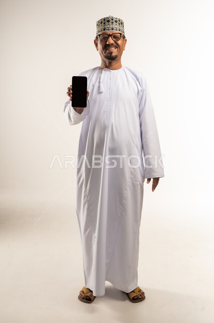 Showing a blank black mobile screen, using modern technologies and advanced devices, portrait of an Arab Gulf Omani man wearing a dishdasha and a kummah holding a mobile phone in his hand, looking at the camera with gestures of happiness and pleasure, advertisement and promotional propaganda, full-length body, white background