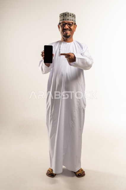 الإشارة إلى شاشة  جوال سوداء فارغة، استخدام التقنيات الحديثة والأجهزة المتطورة، بورتريه لرجل عربي خليجي عماني يرتدي الدشداشة والكمة يمسك بيده الهاتف المحمول، النظر إلى الكاميرا بإيماءات السعادة والسرور، إعلان ودعاية ترويجية، الجسم بالطول الكامل، خلفية بيضاء