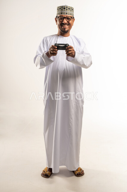 لعب ألعاب الفيديو بإيماءات السرور والسعادة، المتعة والتسلية والتفاعل، قضاء وقت ممتع وترفيهي في استخدام التكنولوجيا الحديثة، بورتريه لرجل عربي خليجي عماني يرتدي الدشداشة والكمه يمسك بعصا التحكم بتعابير النصر والحماس، الجسم بالطول الكامل، خلفية بيضاء