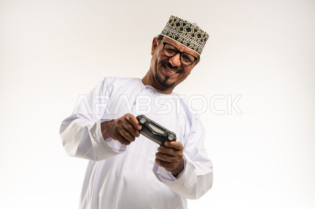 أنشطة ترفيهية منزلية، لعب ألعاب الفيديو بإيماءات السرور والسعادة، المتعة والتسلية والتفاعل، قضاء وقت ممتع وترفيهي في استخدام التكنولوجيا الحديثة ، بورتريه مقرب لرجل عربي خليجي عماني يرتدي الدشداشة والكمه يمسك بعصا التحكم بتعابير النصر والحماس، خلفية بيضاء