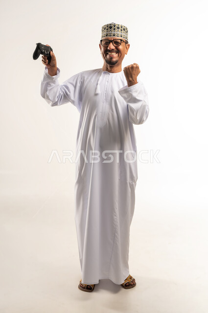 المتعة والتسلية والتفاعل، قضاء وقت ممتع وترفيهي في استخدام التكنولوجيا الحديثة ، بورتريه لرجل عربي خليجي عماني يرتدي الدشداشة والكمه يمسك بعصا التحكم يرفع قبضته بتعابير النصر والحماس، لعب العاب الفيديو بإيماءات السرور والسعادة، الجسم بالطول الكامل، خلفية بيضاء