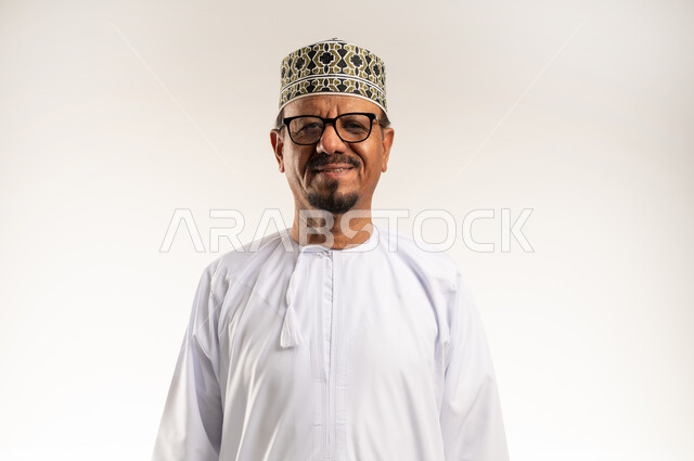 الأناقة والاهتمام بالمظهر الخارجي, أزياء و ملابس خليجية عمانية, بورتريه مقرب لرجل عربي خليجي عماني يرتدي الدشداشه و الكمه يلبس النظارات الطبية وينظر الى الكاميرا بإيماءات السعادة و السرور, خلفية بيضاء