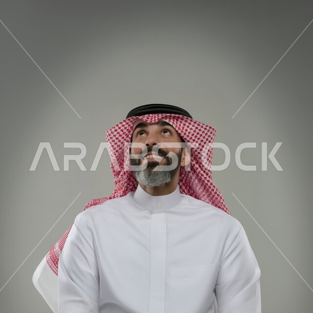 الاهتمام بالمظهر الخارجي، مفهوم الأناقة والرجولة، بورتريه مقرب لرجل عربي خليجي سعودي مبتسم يرتدي الثوب والشماغ التقليدي ينظر إلى الاعلى بإيماءات السرور والسعادة، خلفية رمادية