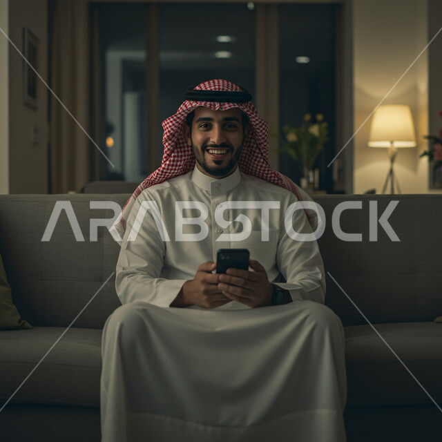 التواصل والدردشة مع الأصدقاء والأقارب عبر الجوال، ايماءات الفرح والسعادة، رجل عربي خليجي سعودي يرتدي الثوب التقليدي والشماغ يجلس في غرفة المعيشة يمسك بيده الهاتف المحمول، قضاء وقت ممتع في المنزل، استخدام جهاز تقني حديث ومتطور