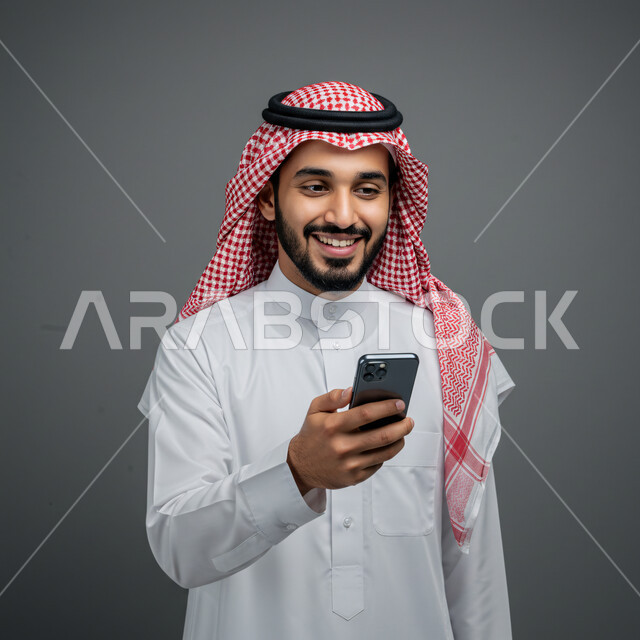 الدردشة مع الأصدقاء والأقارب عبر الجوال، تصفح مواقع التواصل الاجتماعي، استخدام جهاز تقني حديث ومتطور، بورتريه مقرب لرجل عربي خليجي سعودي يرتدي الثوب التقليدي والشماغ يمسك بيده الهاتف المحمول، خلفية رمادية