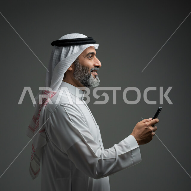 تصفح مواقع التواصل الاجتماعي، الدردشة مع الأصدقاء والأقارب عبر الجوال، استخدام جهاز تقني حديث ومتطور، بورتريه مقرب من الجانب لرجل عربي خليجي سعودي يرتدي الثوب التقليدي والشماغ يمسك بيده الهاتف المحمول، خلفية رمادية