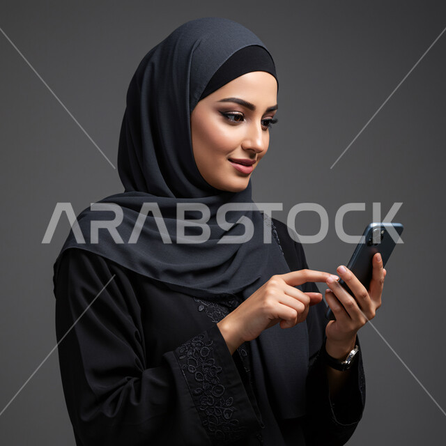 Browsing social networking sites, using a modern and advanced technical device, chatting with friends and relatives via mobile, close-up portrait of a veiled Saudi Arabian Gulf woman wearing an abaya holding a mobile phone in her hand, gray background