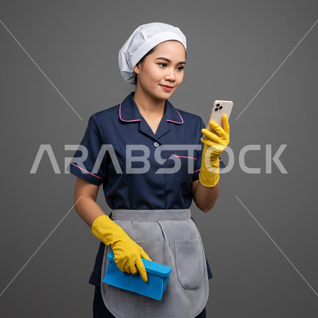 تصفح مواقع التواصل الاجتماعي، استقطاب العاملات من الخارج، الدردشة مع الأصدقاء والأقارب عبر الجوال، استخدام جهاز تقني حديث ومتطور، بورتريه لعاملة اجنبية ترتدي اليونيفورم تمسك بيدها الهاتف المحمول ادوات التنظيف، خلفية رمادية