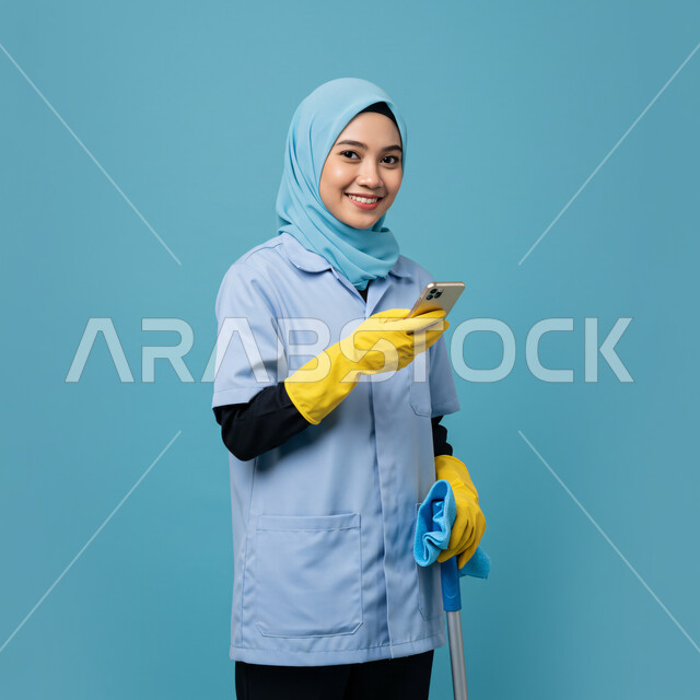 Chatting with friends and relatives via mobile phone, recruiting female workers from abroad, browsing social networking sites, using a modern and advanced technical device, portrait of a foreign female worker wearing a uniform holding a mobile phone and cleaning tools in her hand, blue background