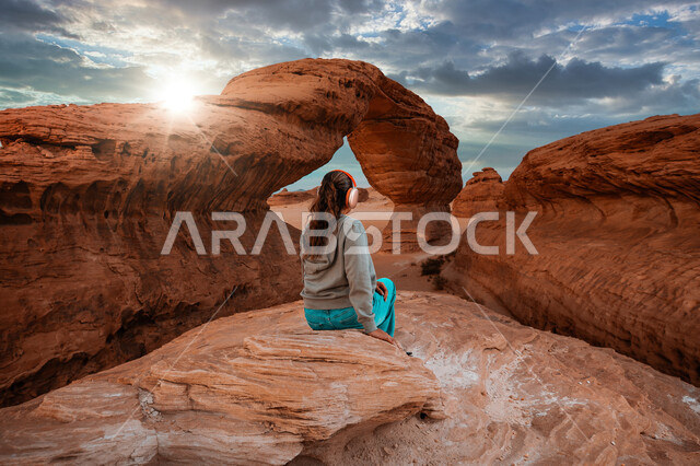 معالم أثرية تاريخية قديمة، صورة من الخلف لسائحة ترتدي الزي الكاجوال تجلس أمام صخرة القوس الشهيرة في المملكة العربيه السعودية، اماكن سياحية طبيعية مشهورة، التكوينات والتشكيلات الصخرية في صحراء محافظة العلا، القمم والمرتفعات الجبلية وسط البيئة الرملية الصحراوية