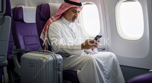 الدردشة مع الأهل والأصدقاء عبر الموبايل, خطوط الطيران السعودية، تصفح الانترنت ومواقع التواصل الاجتماعي، رحلات جوية سياحية، صورة من الجانب لرجل عربي خليجي سعودي يرتدي الثوب التقليدي والشماغ يجلس في الطائرة بجانب حقيبة السفر يحمل بيده الهاتف المحمول