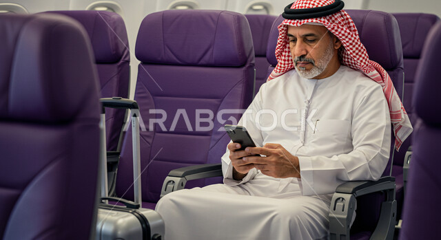 رحلات جوية سياحية، تصفح الانترنت ومواقع التواصل الاجتماعي، الدردشة مع الأهل والأصدقاء عبر الموبايل, رجل عربي خليجي سعودي يرتدي الثوب التقليدي والشماغ يجلس في الطائرة بجانب حقيبة السفر يحمل بيده الهاتف المحمول، خطوط الطيران السعودية
