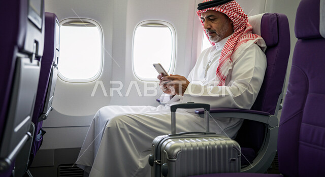 الدردشة مع الأهل والأصدقاء عبر الموبايل, خطوط الطيران السعودية، تصفح الانترنت ومواقع التواصل الاجتماعي، رحلات جوية سياحية، صورة من الجانب لرجل عربي خليجي سعودي يرتدي الثوب التقليدي والشماغ يجلس في الطائرة بجانب حقيبة السفر يحمل بيده الهاتف المحمول