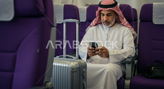 رحلات جوية سياحية، تصفح الانترنت ومواقع التواصل الاجتماعي، الدردشة مع الأهل والأصدقاء عبر الموبايل, رجل عربي خليجي سعودي يرتدي الثوب التقليدي والشماغ يجلس في الطائرة بجانب حقيبة السفر يحمل بيده الهاتف المحمول، خطوط الطيران السعودية