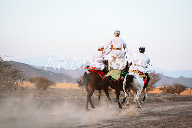Wandering through the desert on horseback in the Bahla region, the passion for equestrianism and the hobby of horse riding, the relationship of the people of the country with the purebred Omani horse, a picture from the back of Arab Gulf Omani men wearing the dishdasha and the kuma riding the backs of horses, following the traditions of the ancestors in caring for and raising horses