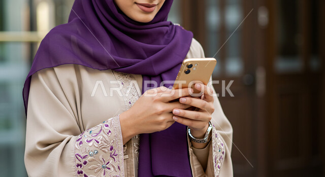 الدردشة مع الاهل والاصدقاء، تصفح الانترنت ومواقع التواصل الاجتماعي، استخدام جهاز تقني حديث ومتطور، بورتريه مقرب لامرأة عربية خليجية سعودية محجبة مبتسمة ترتدي العباءة تحمل بيدها الهاتف المحمول