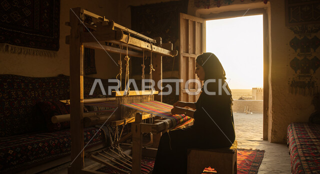 الفن التراثي الحرفي التقليدي، نقش وزخرفة من التراث السعودي، معمل ومصنع لحياكة وغزل السجاد، مهنة وحرفة شعبية، صورة من الجانب لسيدة عربية خليجية سعودية محجبة ترتدي العباءة تقوم بنسج السجادة، منتج صناعي محلي وطني