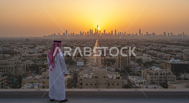 النمو و التطور العمراني في المدينة، الاعتزاز والفخر بالتقدم المعماري، التجول والتنزه واستنشاق الهواء النقي في الخارج، صورة من الخلف لرجل عربي خليجي سعودي يرتدي الشماغ و الثوب التقليدي يقوم بمشاهدة غروب الشمس