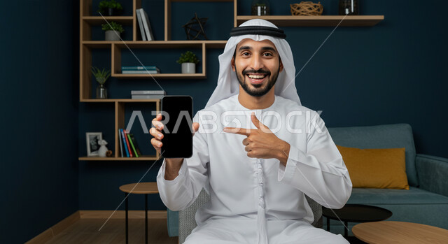 ايماءات السعادة والسرور، استخدام جهاز تقني حديث ومتطور، عرض شاشة سوداء فارغة عبر الهاتف المحمول، صورة مقربة لرجل عربي خليجي اماراتي مبتسم يرتدي الكندورة والغتره يجلس في غرفة المعيشة ويقوم بالاشارة الى الجوال باصبع السبابة وينظر إلى الكاميرا
