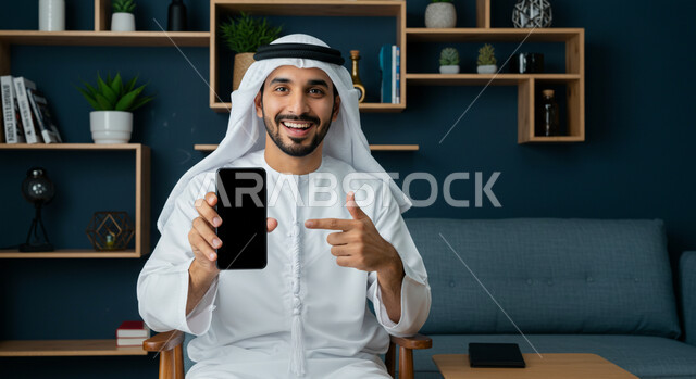 Using a modern and advanced technical device, gestures of happiness and pleasure, displaying a blank black screen via a mobile phone, a close-up image of a smiling Emirati Gulf Arab man wearing a kandura and a ghutra sitting in the living room and pointing at the mobile phone with his index finger and looking at the camera