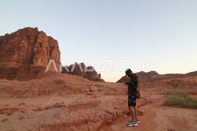 التكوينات والتشكيلات الصخرية على الوديان الجبلية في الأماكن الأثرية التاريخية، أرض النخيل والعيون، مكان ومنطقة سياحية مشهورة، التضاريس الجبلية الطبيعية في المملكة العربية السعودية، سائح أجنبي يمسك بيده هاتف محمول يقف في وادي الديسة في منطقة تبوك في النهار