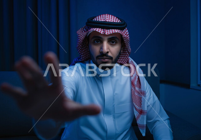 إيماءات الحماس والاندماج والتركيز, التفاعل ومحاكاة العناصر الخيالية, محاولة إمساك شيء ما بحركات الدهشة، صورة مقربة لشاب عربي خليجي سعودي يرتدي الثوب التقليدي والشماغ يجلس على كرسي ويشير بيده إلى الأمام 