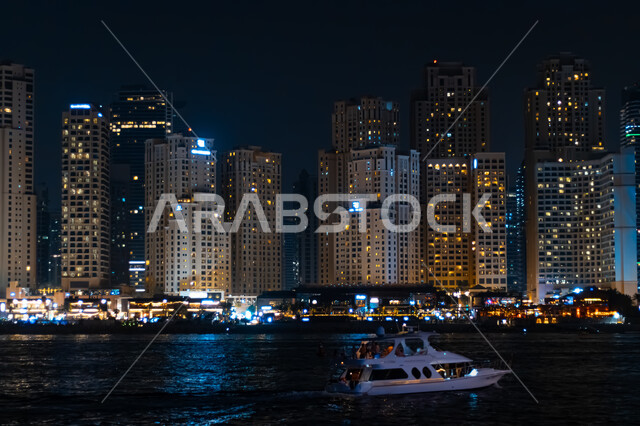 الفن الهندسي المعماري لبناء الأبراج وناطحات السحاب على الطراز الحديث, مفهوم التقدم والتطور العمراني, مكان سياحي مشهور في الإمارات العربية المتحدة, المباني والمنشآت المضيئة في الليل بمدينة دبي, قارب سياحي يسير في البحر