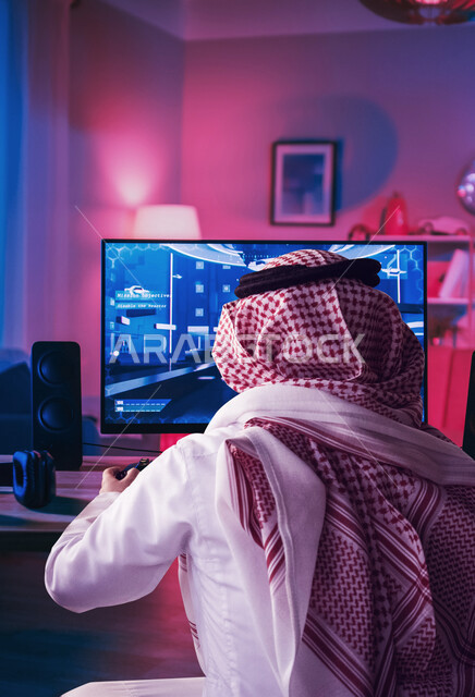 صورة بزاوية من الخلف لشاب عربي خليجي سعودي يلعب ألعاب الفيديو على جهاز الكمبيوتر ، يحمل يد التحكم ويلعب ألعاب الفيديو ، التسليه والترفيه ، قضاء أوقات ممتعة وقت الفراغ ، التحدي والتركيز ، مفهوم إدمان الإنترنت والأجهزة الإلكترونية الحديثة