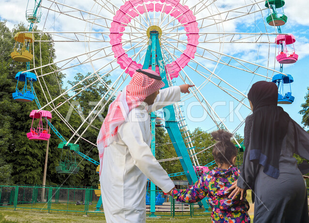 صورة بزاوية من الخلف لعائلة عربية خليجية سعودية يقضون اوقاتهم بالمرح والتسلية في مدينة الألعاب في وضع النهار ، قضاء اوقات عائلية ممتعة في مدينة الملاهي ، التسلية والترفيه ، الترفيه في العطلة الصيفية ، عطلة نهاية الأسبوع ، عجلة فيريس