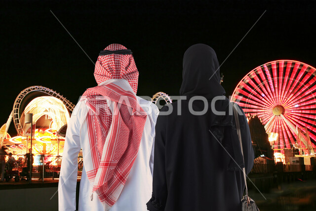 صورة بزاوية من الخلف لزوجان عربيان خليجيان سعوديان لحظة دخولهما مدينة الألعاب ، قضاء اوقات عائلية ممتعة في مدينة الملاهي في وضع االليل ، التسلية والترفيه ، الترفيه في العطلة الصيفية ، عطلة نهاية الأسبوع