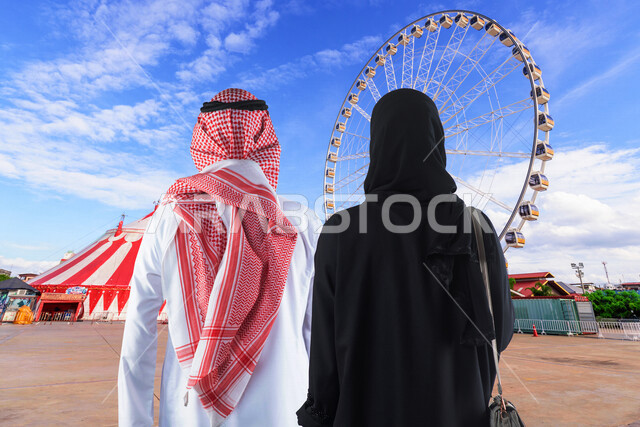 صورة بزاوية من الخلف لزوجان عربيان خليجيان سعوديان لحظة دخولهما مدينة الألعاب ، قضاء اوقات عائلية ممتعة في مدينة الملاهي في وضع النهار ، التسلية والترفيه ، الترفيه في العطلة الصيفية ، عطلة نهاية الأسبوع