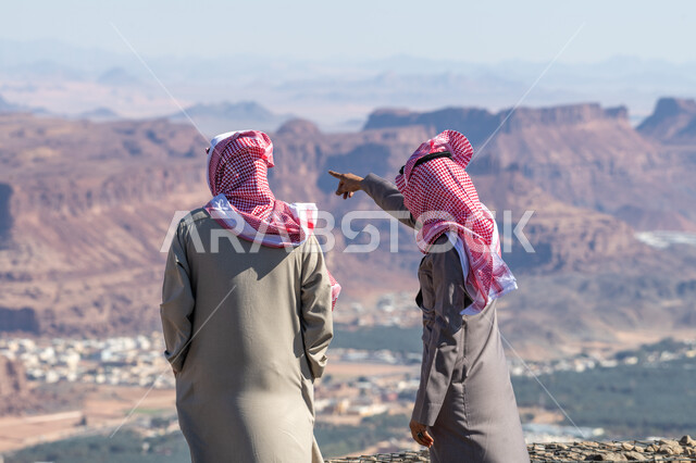 Mountain peaks and heights, tourist attractions to attract and attract tourists, a picture from the back of two Saudi Arabian Gulf men wearing traditional thobe and shemagh standing in front of historical monuments in the city of Al-Ula, natural rock formations and formations in the Kingdom of Saudi Arabia