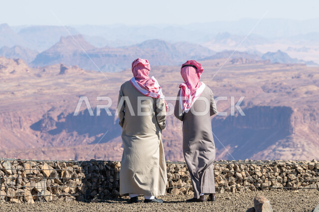 القمم والمرتفعات الجبلية، أماكن سياحية لجذب واستقطاب السياح، صورة من الخلف لرجلين عربيين خليجيين سعوديين يرتديان الثوب التقليدي والشماغ يقفان أمام المعالم التاريخية الأثرية في مدينة العلا، التكوينات والتشكيلات الصخرية الطبيعية بالمملكة العربية السعودية