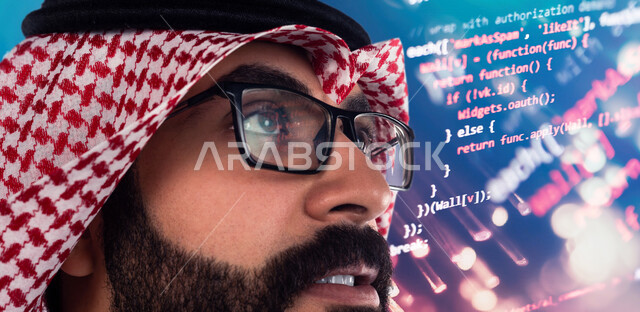 صورة مقربة لوجه رجل عربي خليجي سعودي يقف بجانب شاشة تحتوي على لغة خاصة بالبرمجة بإيماءات تدل على التركيز و التطلع ، مواكبة الحداثة و التطور التكنولوجي