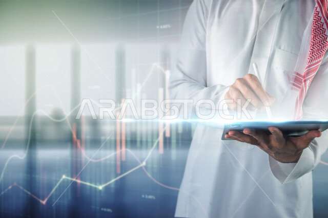 صورة مقربة لرجل عربي سعودي خليجي يحمل بيده قلم يكتب على جهاز تقني ذكي يحمله بيد الأخرى أمام شاشة برسومات بيانية ثلاثية الأبعاد بتقنية الهولوجرام ، متابعة  صعود و نزول البورصة المالية ، الاستثمار المالي و الاقتصادي ، التطور الإقتصادي