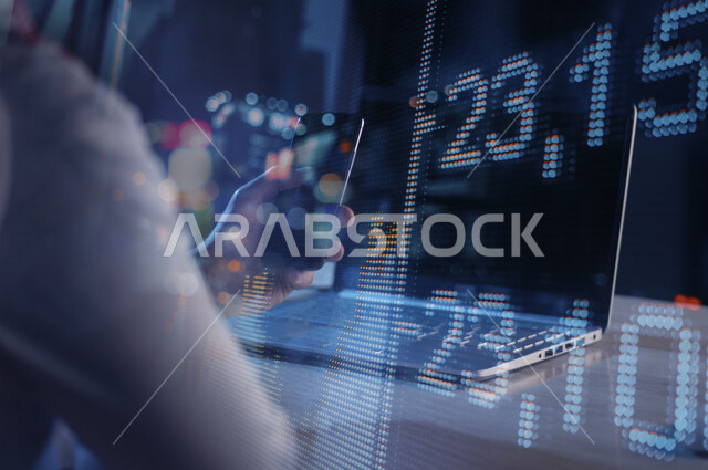 صورة من الخلف لرجل عربي سعودي خليجي يجلس أمام الحاسوب المحمول وبيده الهاتف المحمول يتابع البورصة المالية على شاشة ثلاثية الأبعاد بتقنية الهولوجرام ، صعود و نزول البورصة المالية ، الاستثمار المالي و الاقتصادي ، التطور الإقتصادي