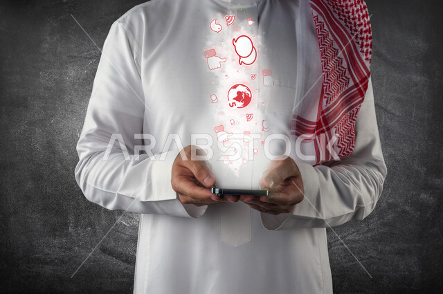 صورة مقربة لرجل عربي خليجي سعودي بين يديه هاتف محمول ، خروج مجموعة متطايرة من الأيقونات الخاصة بتطبيقات الجوال و الأجهزة الإلكترونية بتقنية الهولوجرام ثلاثية الأبعاد