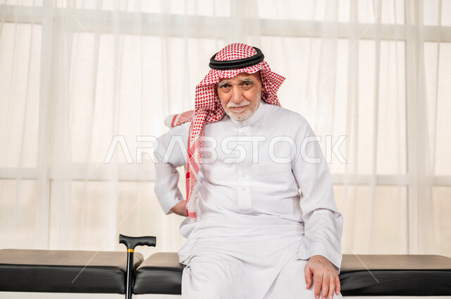 بورتريه رجل عربي سعودي خليجي كبير بالسن يضع يده على ظهره يشعر بحالة من الوجع والألم , صورة مقربة لمسن سعودي يرتدي ثوب أبيض مع شماغ وعقال , مفهوم تعابير الوجه ولغة الجسد , خلفية بيضاء .