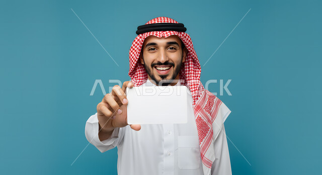 استخدام بطاقة توضيحية تعريفية، بورتريه مقرب لرجل عربي خليجي سعودي مبتسم يرتدي الشماغ والثوب التقليدي يحمل بيده موك أب ينظر إلى الكاميرا بتعابير السرور والسعادة، كروت إعلان وعرض تسويقي، كرت أبيض مستطيل فارغ، مفهوم التسويق لشيء ما، خلفية زرقاء
